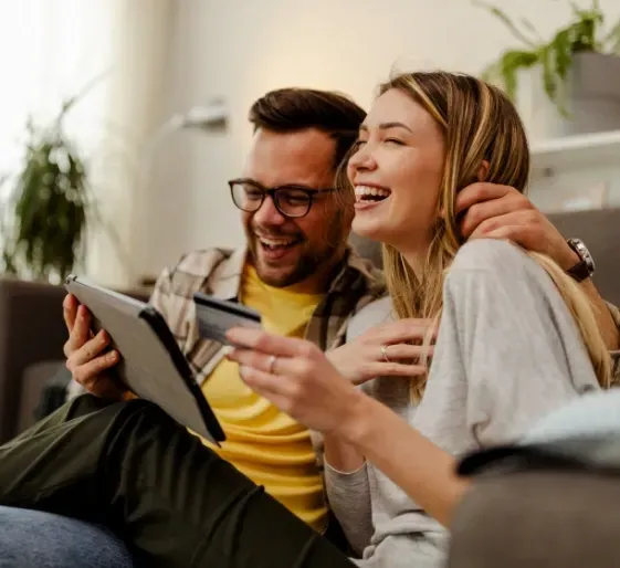 Two people laughing on a couch while looking at a tablet and holding a credit card
