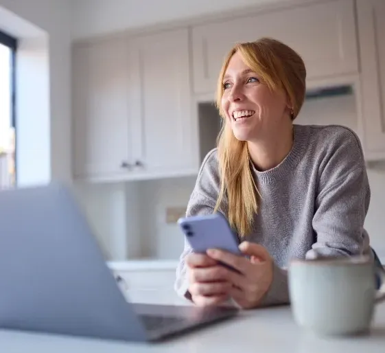 Woman smiling at a kitchen counter, holding a phone beside a laptop and coffee mug