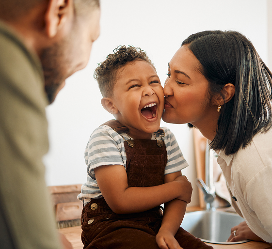 Woman kissing laughing child indoors, with another person blurred in the foreground.