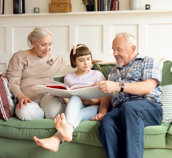 Three people sitting on a green couch reading a book together in a bright living room