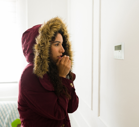 Person in a maroon hooded coat with fur trim, standing indoors by a wall and looking at a thermostat.