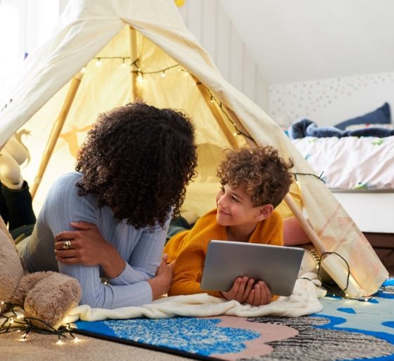 Two children relax in a blanket fort, looking at a laptop on a rug.
