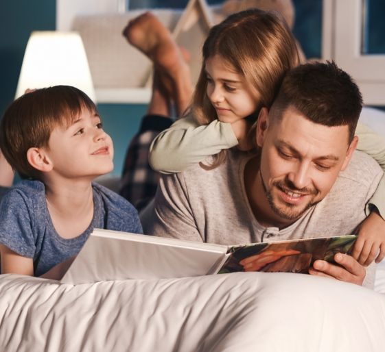 Family reading together in bed, smiling in a cozy bedroom with a lamp nearby.