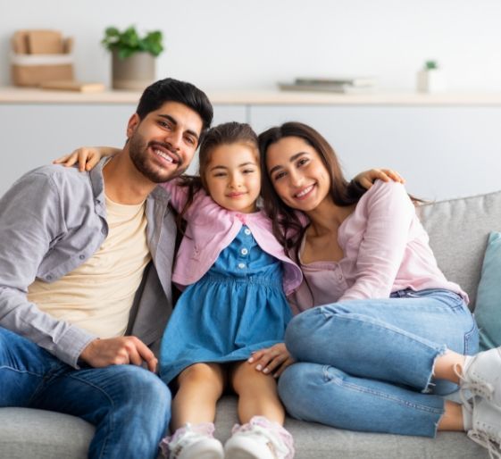 Three people smiling on a couch in a bright living room, with a child in the center wearing a blue dress.