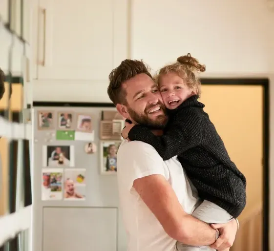 Smiling adult holding a smiling child in a kitchen, with a refrigerator covered in photos in the background