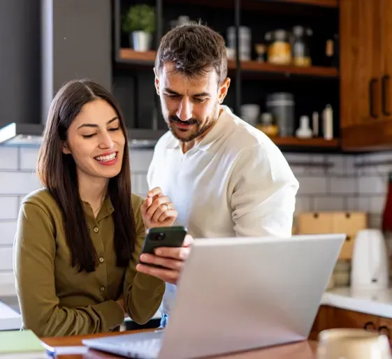 Two people smiling at a laptop in a kitchen, looking at a phone together.