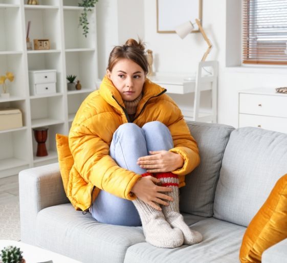 Woman in a yellow puffer jacket sitting curled up on a gray couch in a bright living room.