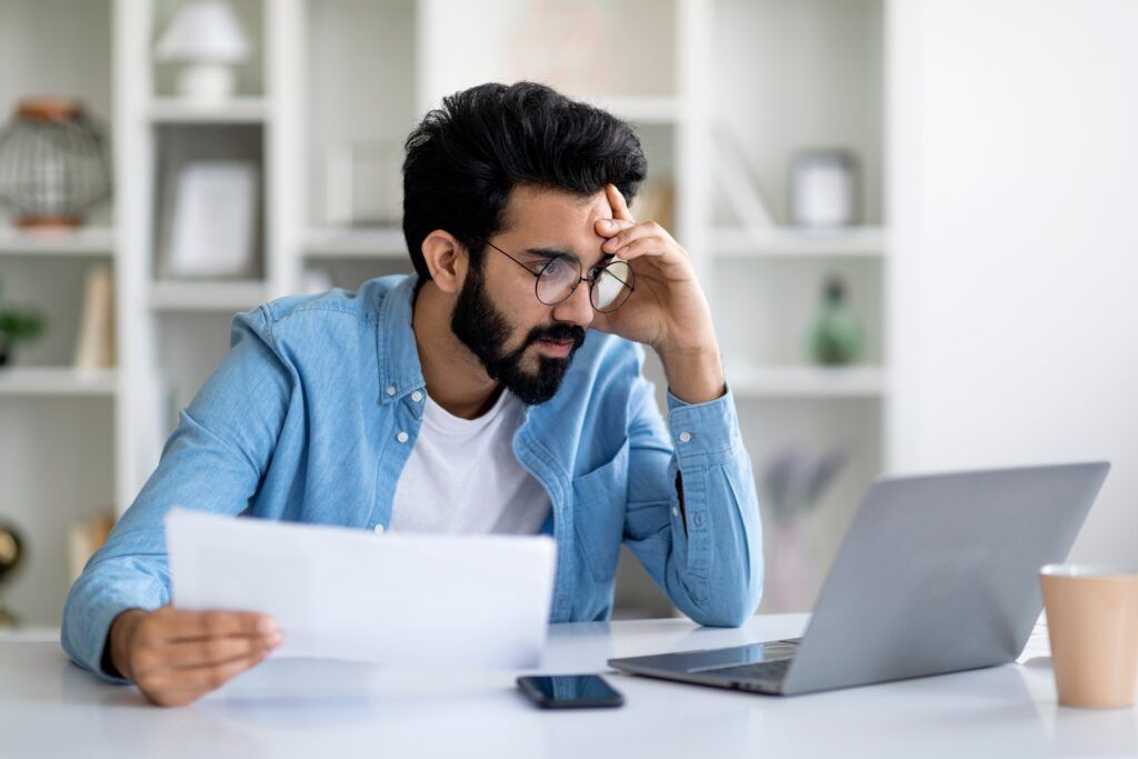 Man at desk reading a paper beside a laptop, looking stressed in a bright home office.