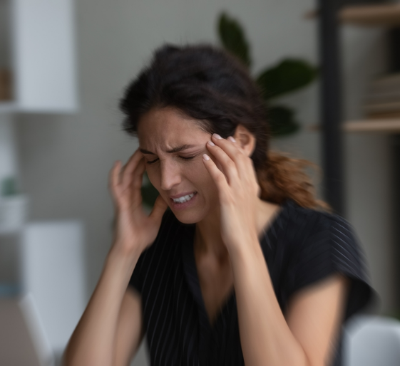 Woman holding her temples with eyes closed, looking stressed in a blurred indoor setting