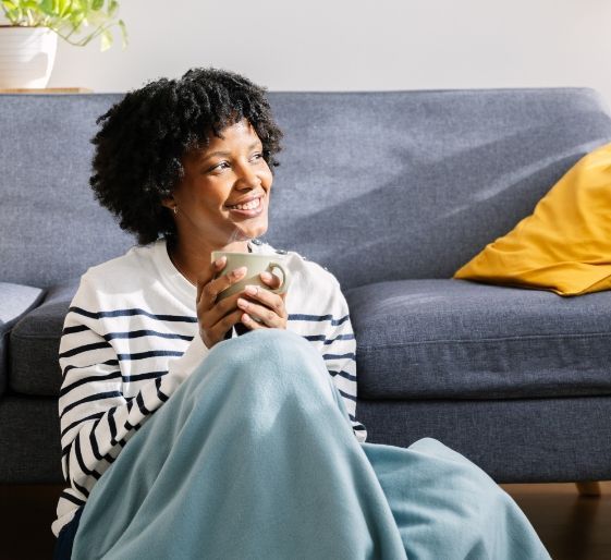 Person sitting on the floor beside a gray sofa, holding a mug and smiling.