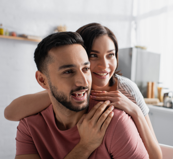 Smiling couple embracing in a bright kitchen, with the woman hugging the man from behind.