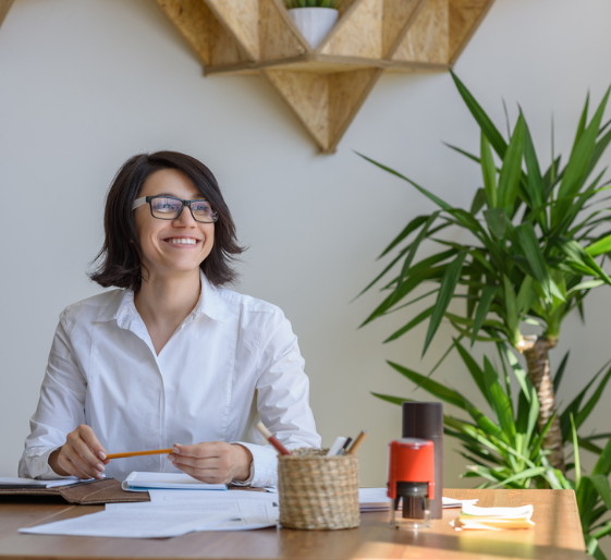 Smiling person in a white shirt at a desk with papers, in a bright office beside a large potted plant