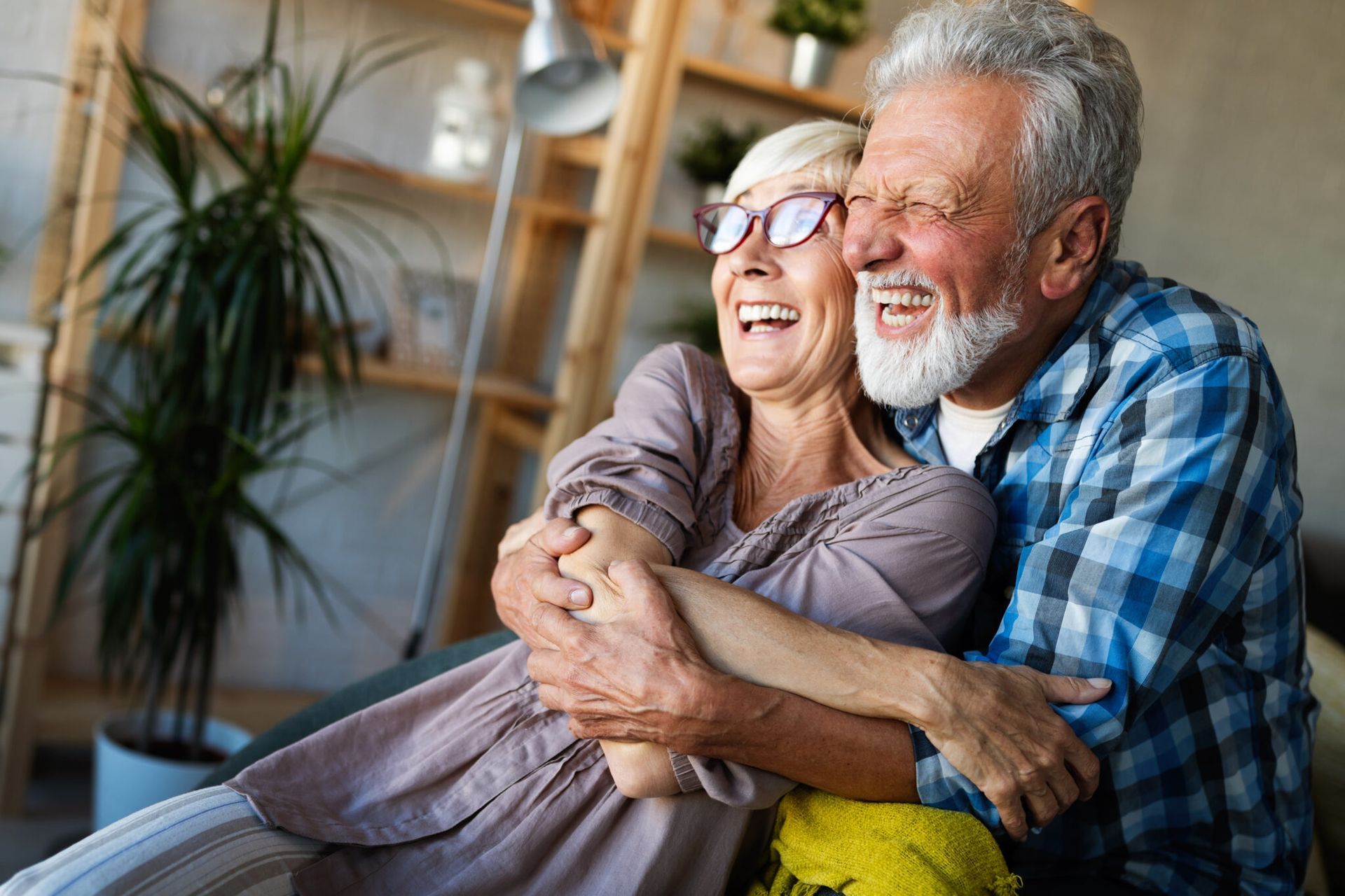 Smiling couple cuddling on a couch in a cozy living room, with a plant and wooden shelves in the background