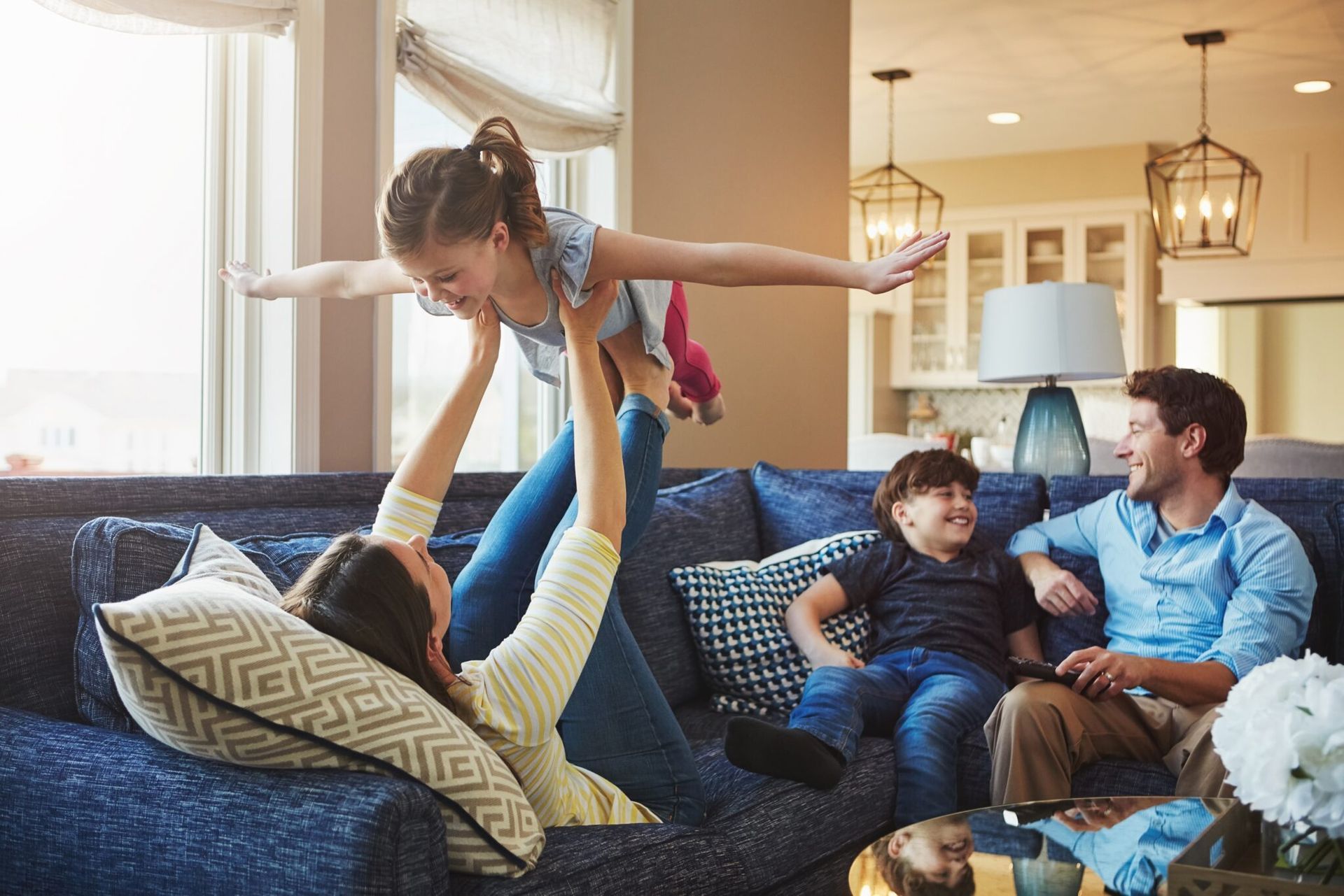 Family playing on a blue couch in a sunlit living room, with a child being lifted playfully above the cushions