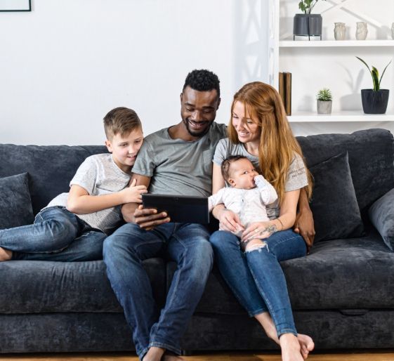 Family sitting on a couch, gathered around a tablet in a bright living room.
