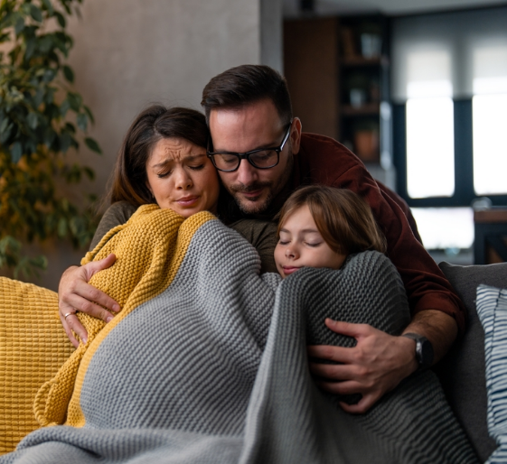 A family cuddles under blankets on a couch, embracing in a cozy living room.