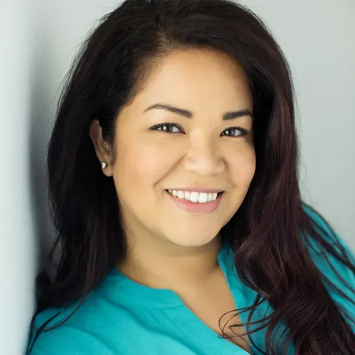 Woman with dark hair and a teal shirt smiles leaning against a white wall.