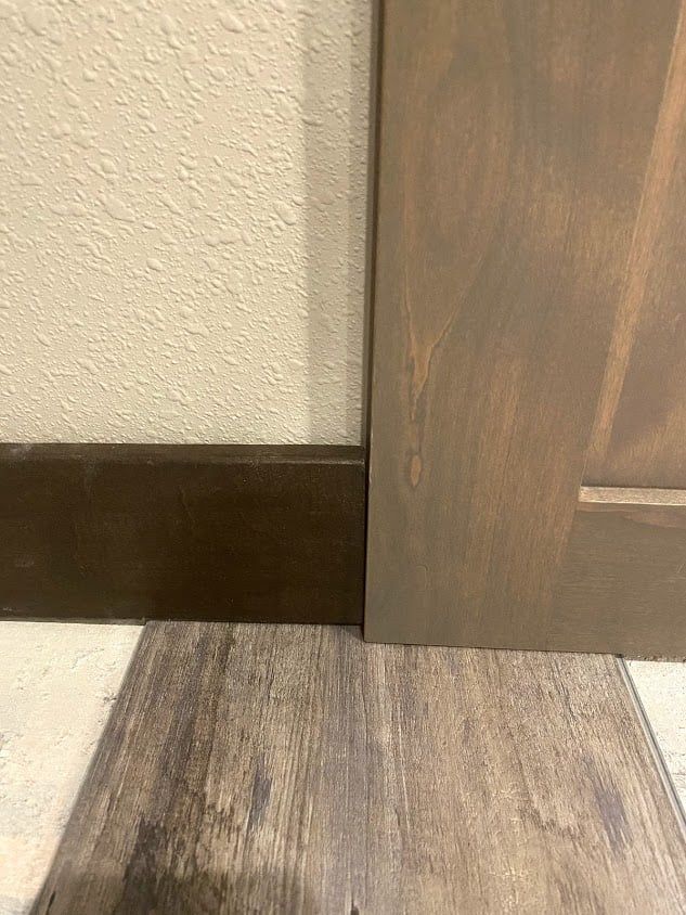Close-up of a wooden door slightly open over a wood floor beside a beige wall and dark baseboard.