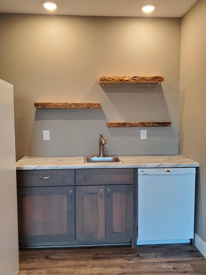 Small kitchenette with sink, wood cabinets, floating shelves, and a mini fridge under recessed lights.