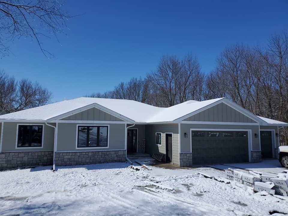 Snow-covered single-story house with gray siding and a two-car garage under a clear blue sky