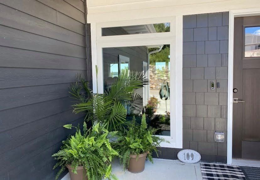 Front porch with potted tropical plants beside a gray house entry and white-framed window