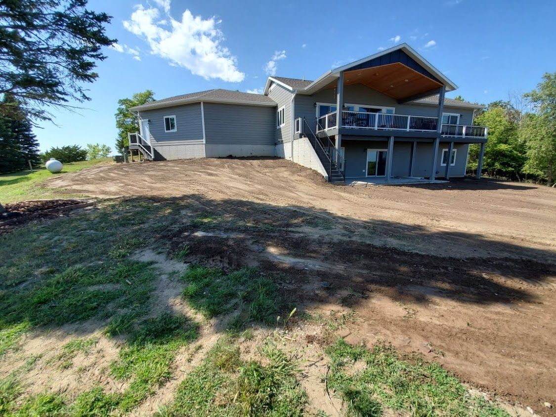 Large gray house with a covered deck on a dirt lot, viewed from the front under a clear blue sky