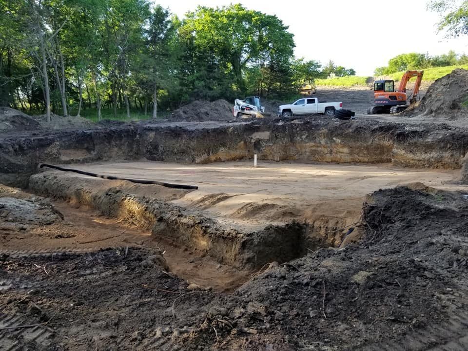Dug-out construction site with muddy earth, a white pickup, and an excavator near trees