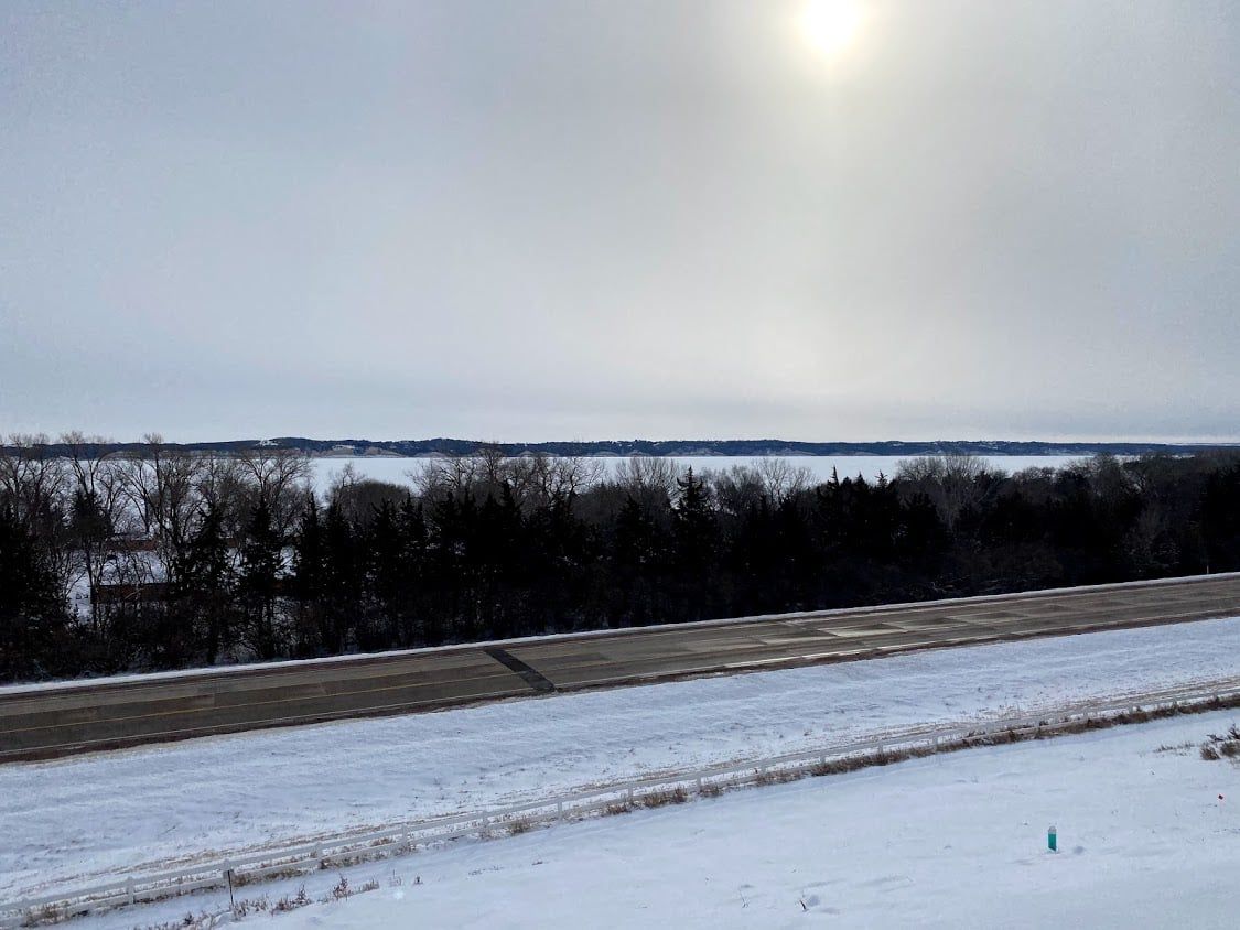 Snowy field and road beside a dark treeline under a pale winter sky with the sun shining through clouds