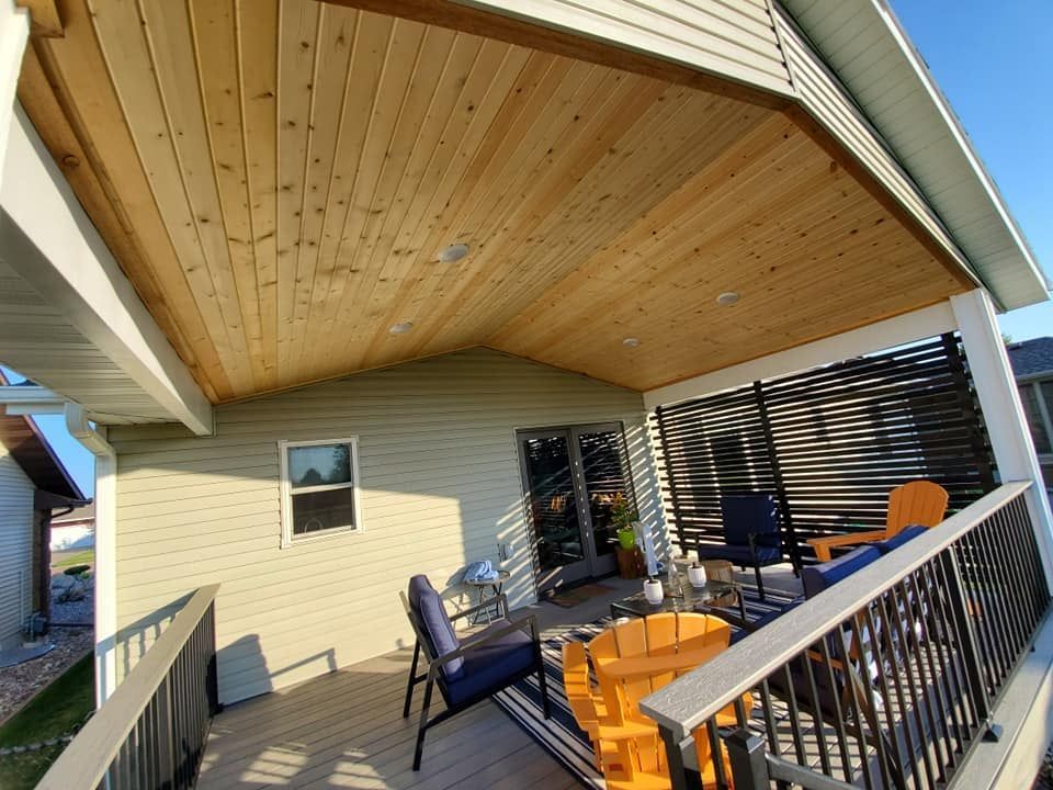 Covered porch with wood ceiling, black railing, and orange chairs outside a beige house.