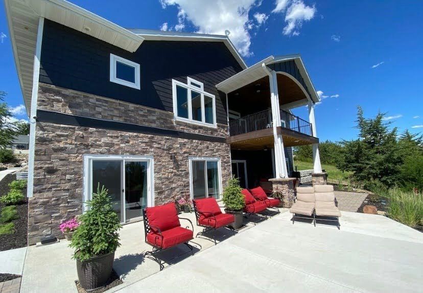 Modern two-story stone house with red chairs and beige patio seating on a sunny deck