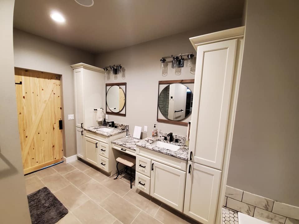 Bright bathroom with double vanity, two mirrors, white cabinets, beige tile floor, and a wooden door.