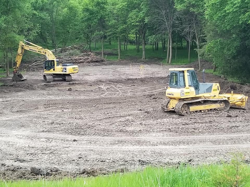 Two yellow excavators working on a dirt clearing surrounded by trees