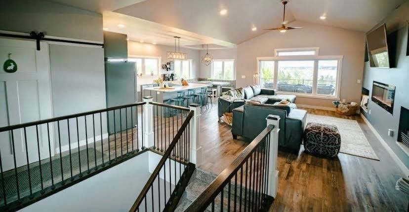 Open-concept living room and kitchen with stairs, wood floors, gray sofa, and large windows overlooking water