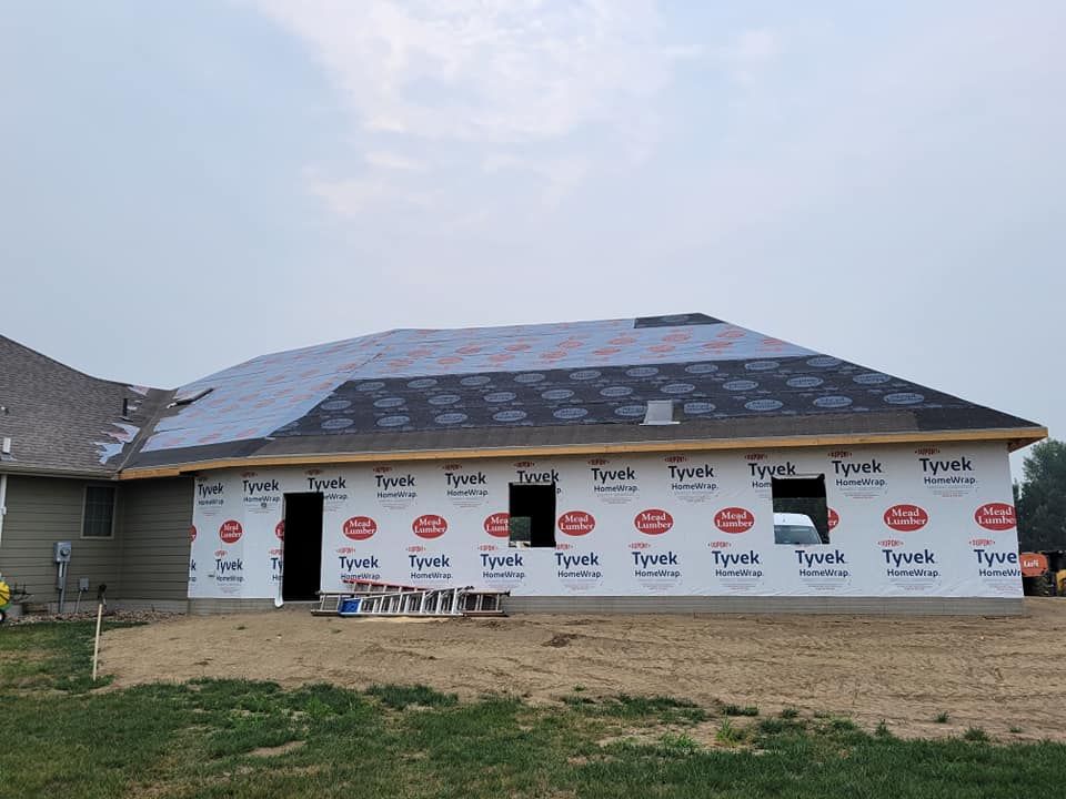 House exterior under construction with roof shingles and white house wrap, viewed from a grassy yard
