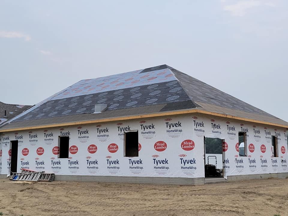 Partially built house wrapped in white Tyvek house wrap with a dark shingled roof on a dirt lot