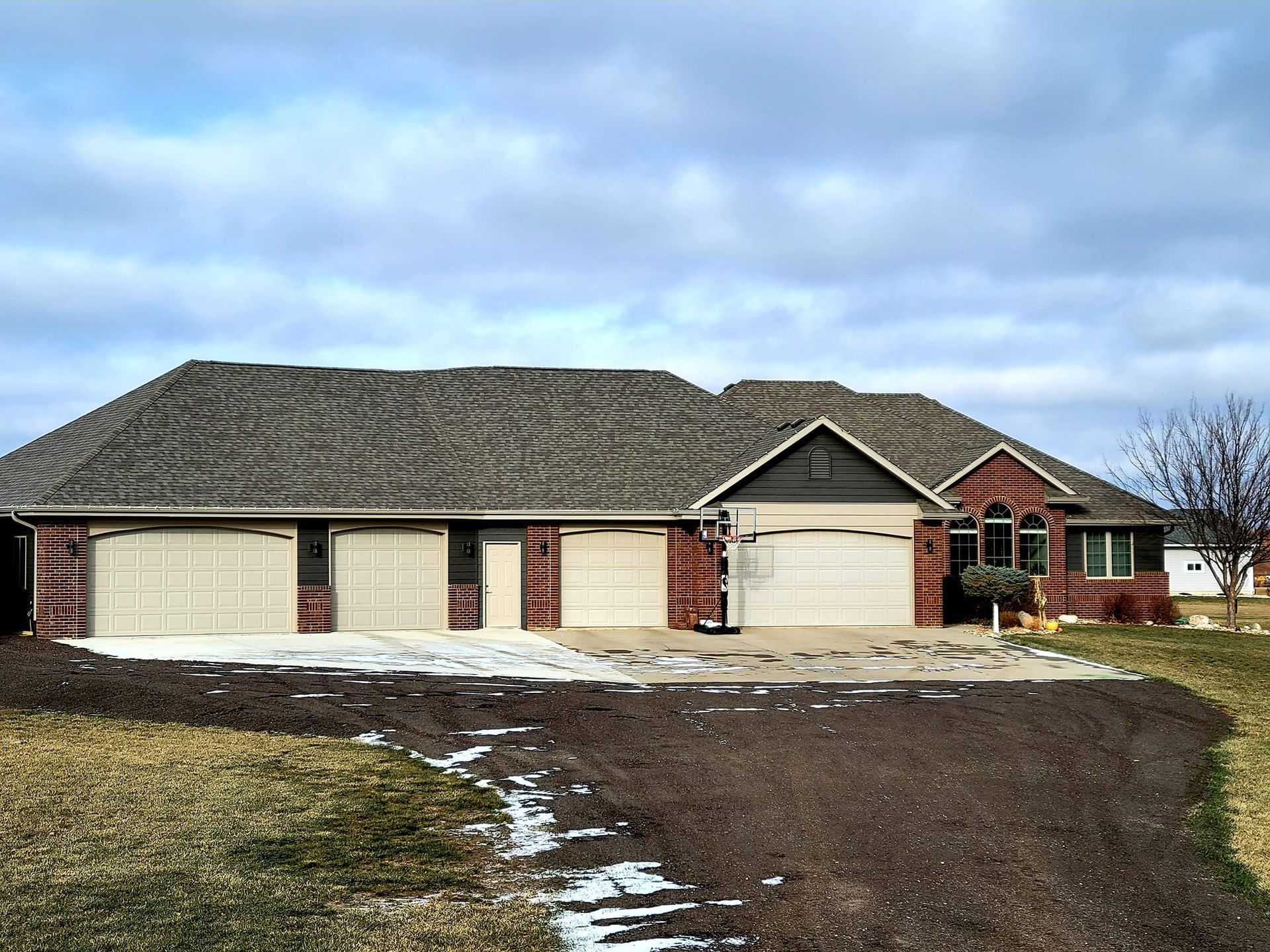 Single-story brick house with two garage doors, a wide driveway, and patches of snow under a cloudy sky