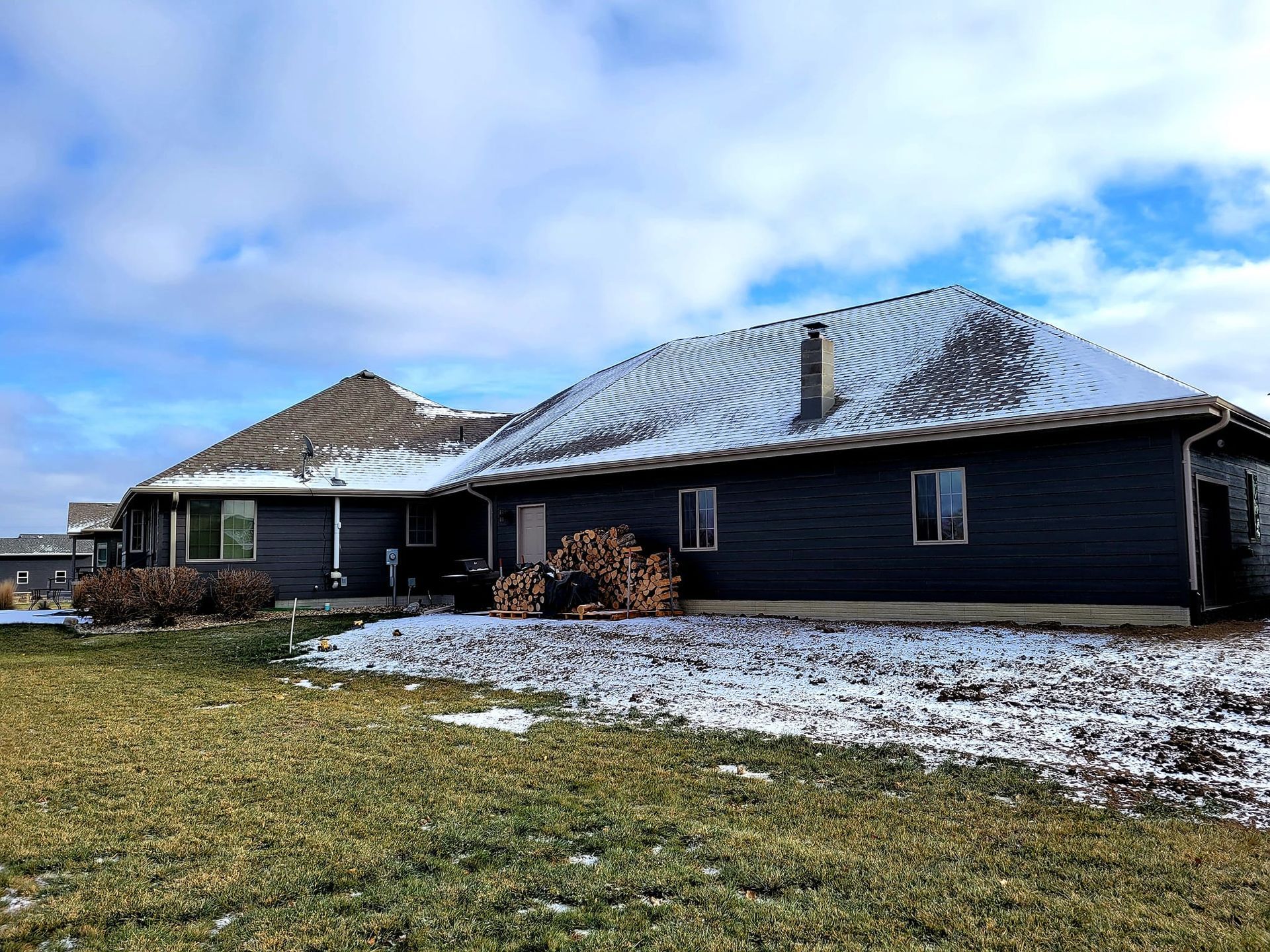 Snow-dusted black house with a dark roof under a cloudy blue sky in a grassy yard