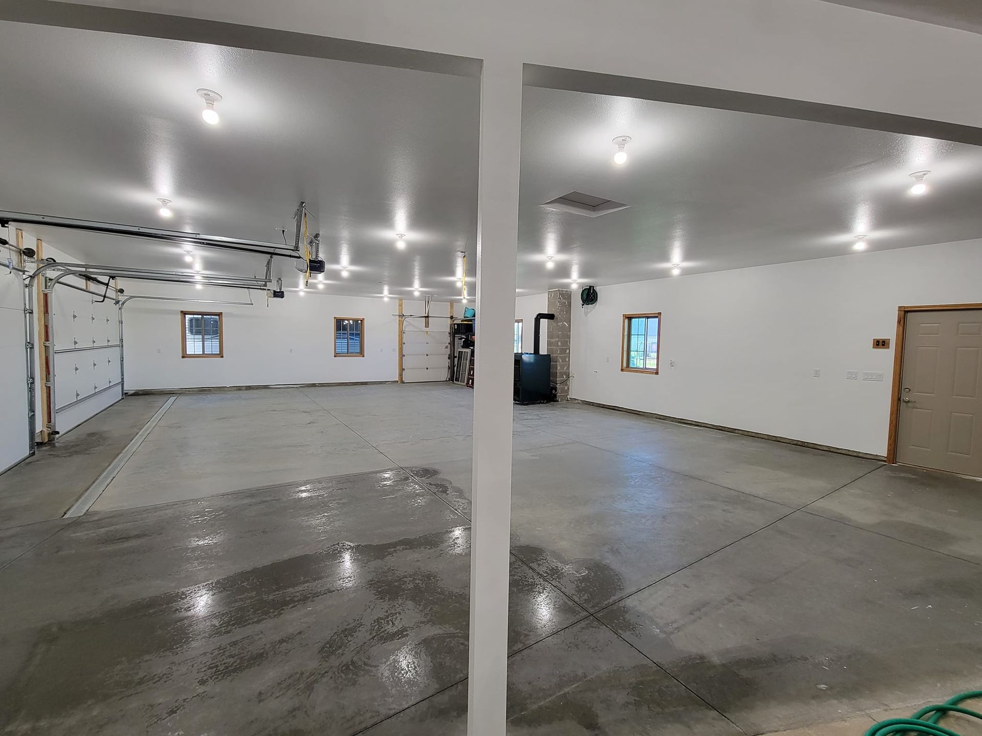 Empty garage interior with white walls, concrete floor, overhead lights, and wet patches on the floor