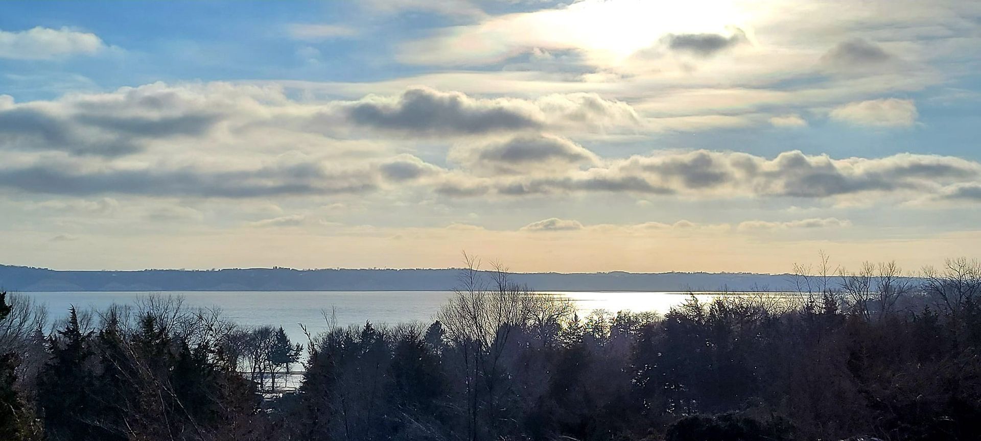 Snowy lakeside landscape with bare trees, calm water, and a cloudy sky glowing with sunlight