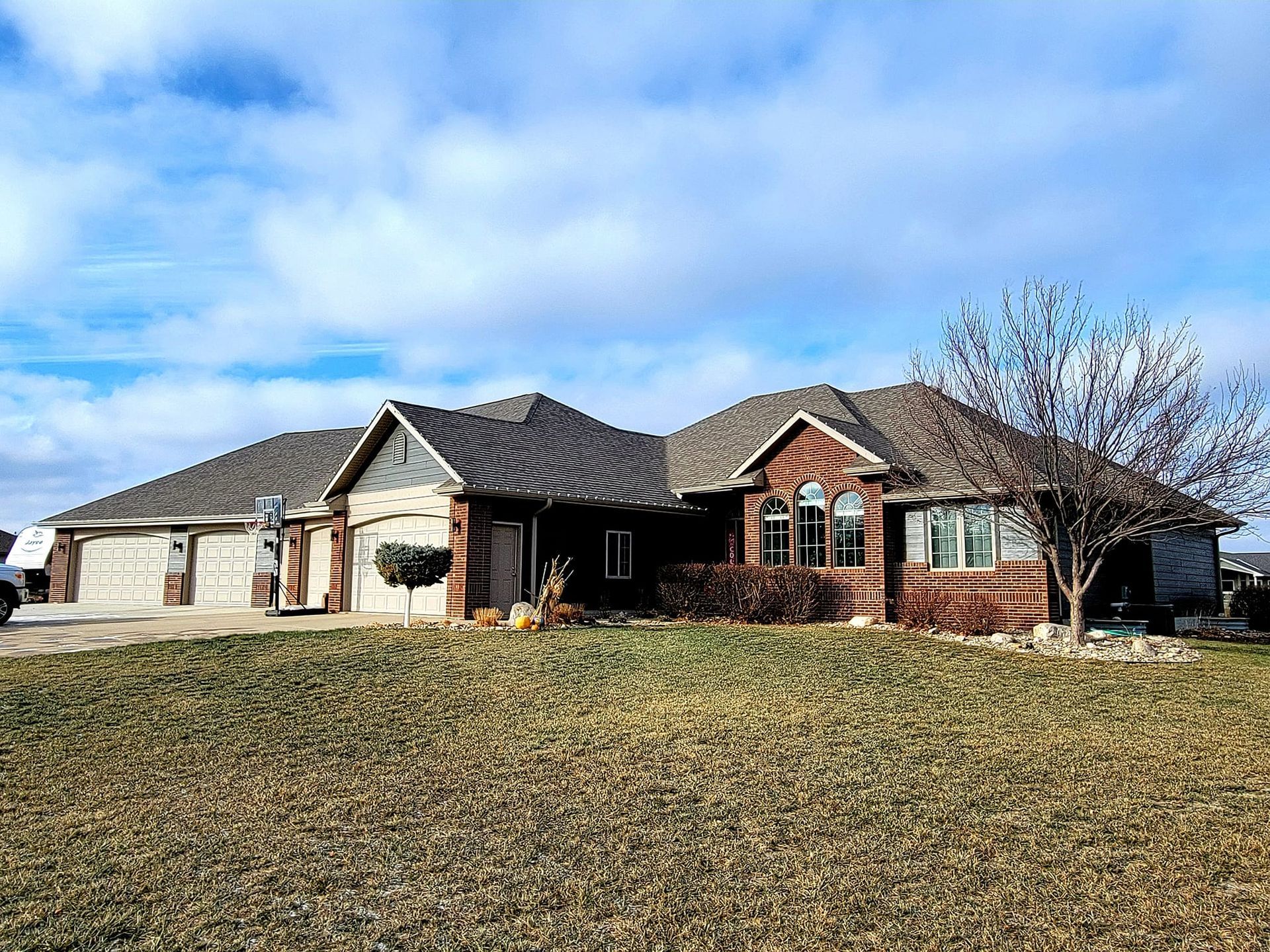 Large brick suburban house with a gray roof and a leaf-covered front lawn under a blue sky