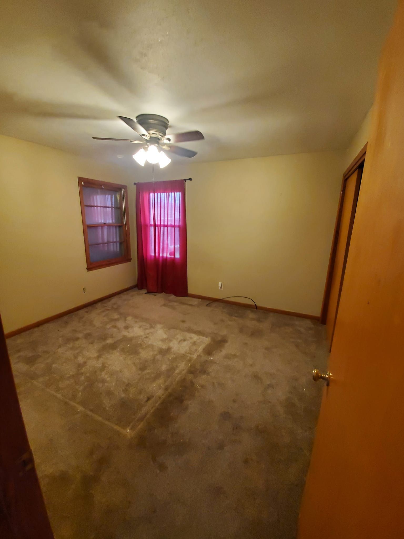 Empty beige bedroom with ceiling fan, brown carpet, and red curtains over two windows.