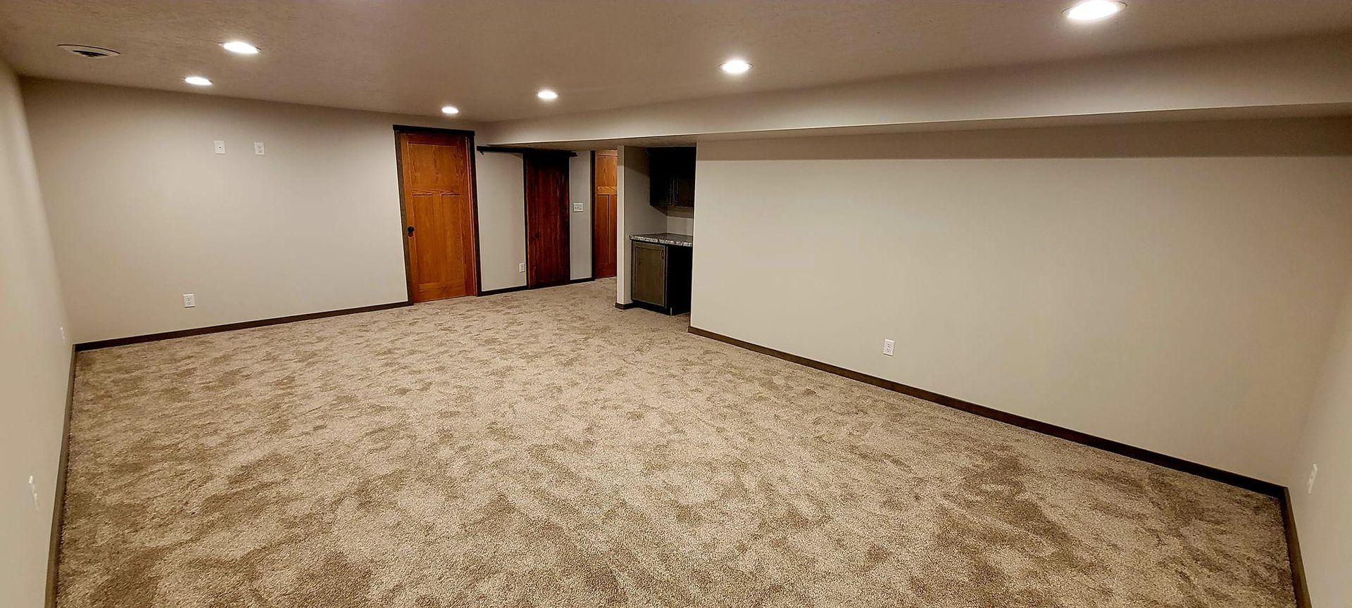 Empty carpeted basement room with recessed lights, white walls, and a doorway near a small cabinet
