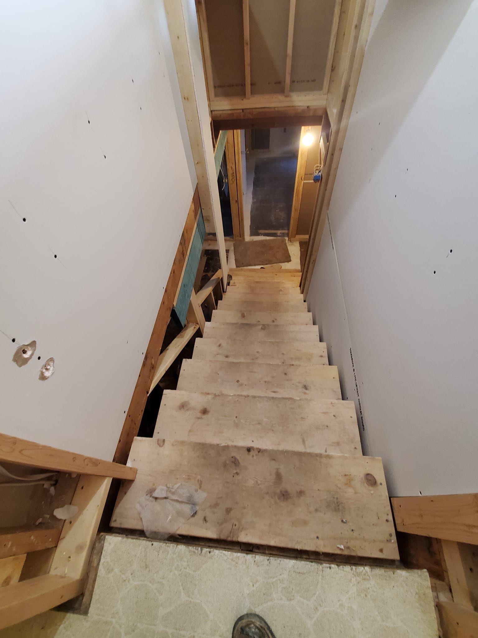 Narrow unfinished stairwell with exposed wood framing and steep stairs leading down to a lit doorway