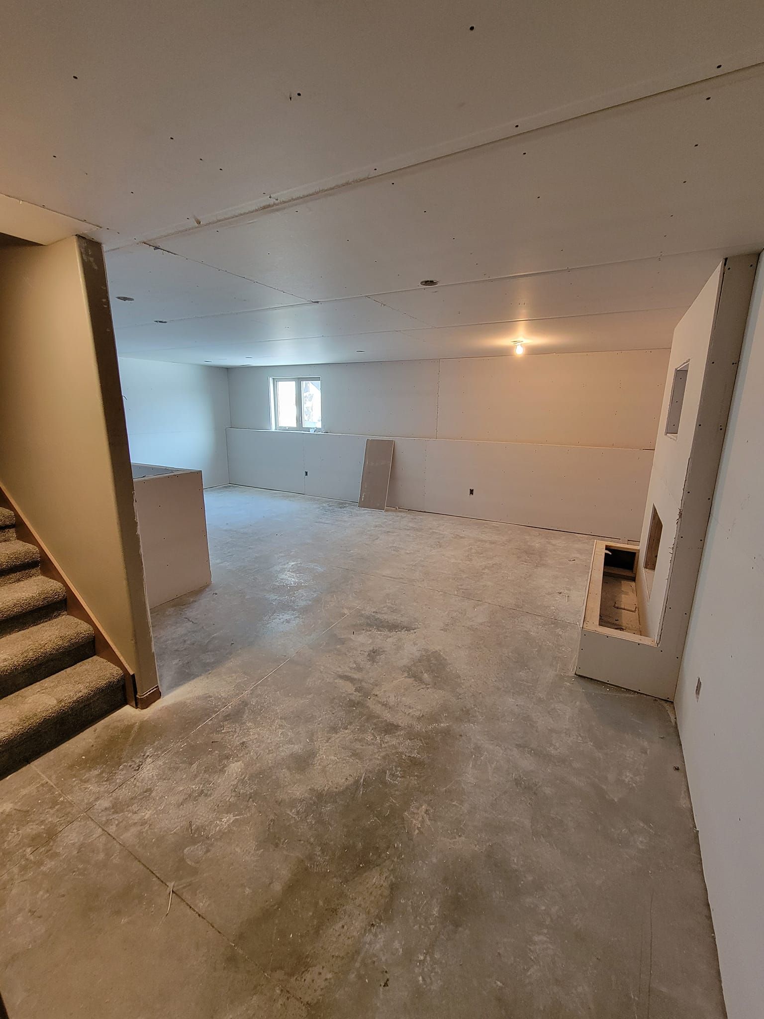 Unfinished basement with concrete floor, staircase, window, and exposed walls lit by a single bulb