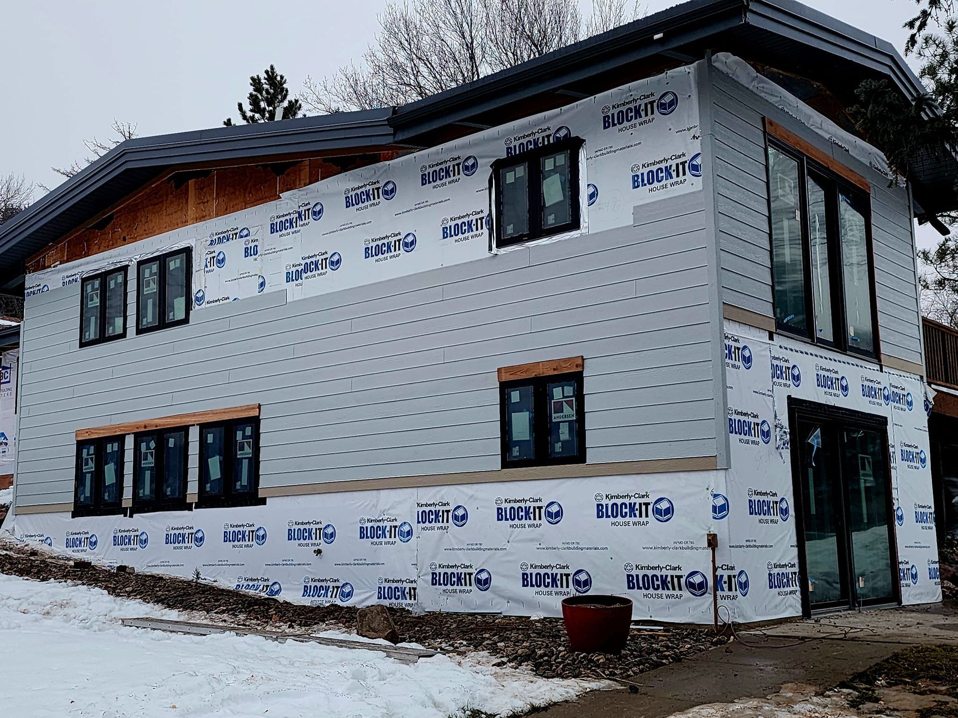 Partially finished house wrapped in white siding under construction, with snow on the ground