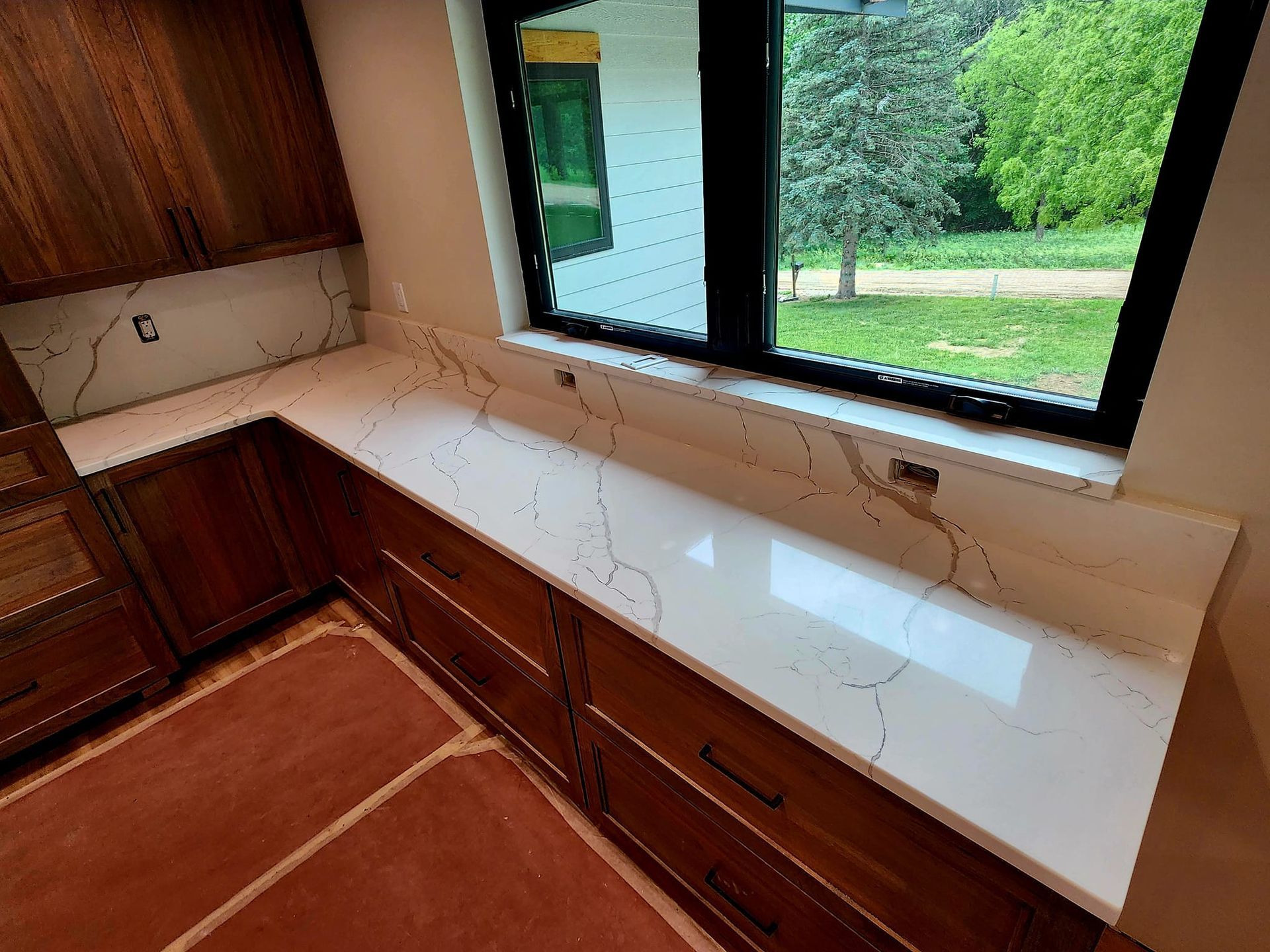 Empty kitchen corner with wooden cabinets, white countertop, and a window overlooking greenery
