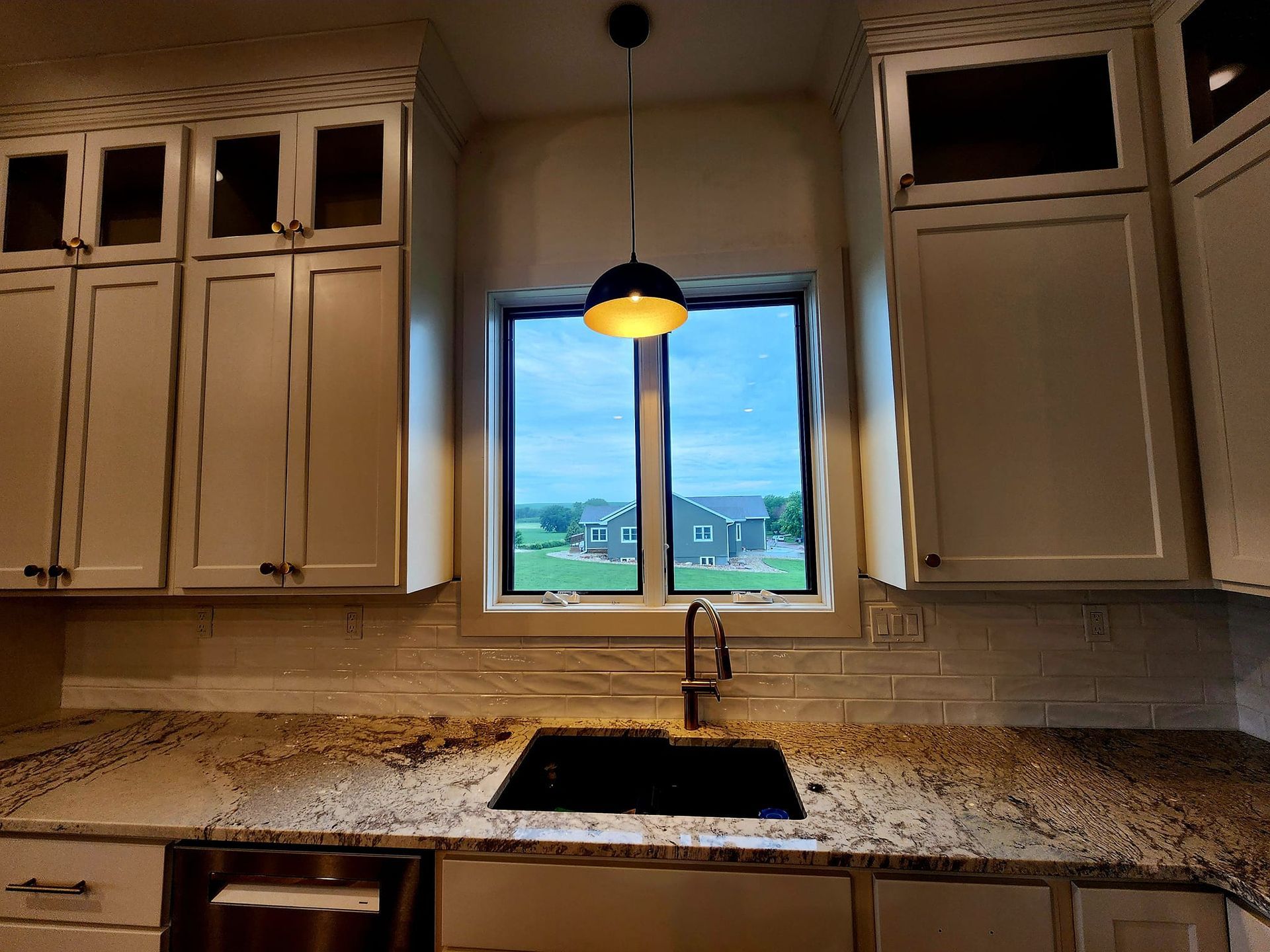 Kitchen sink beneath a window, with beige cabinets, granite countertops, and a pendant light.