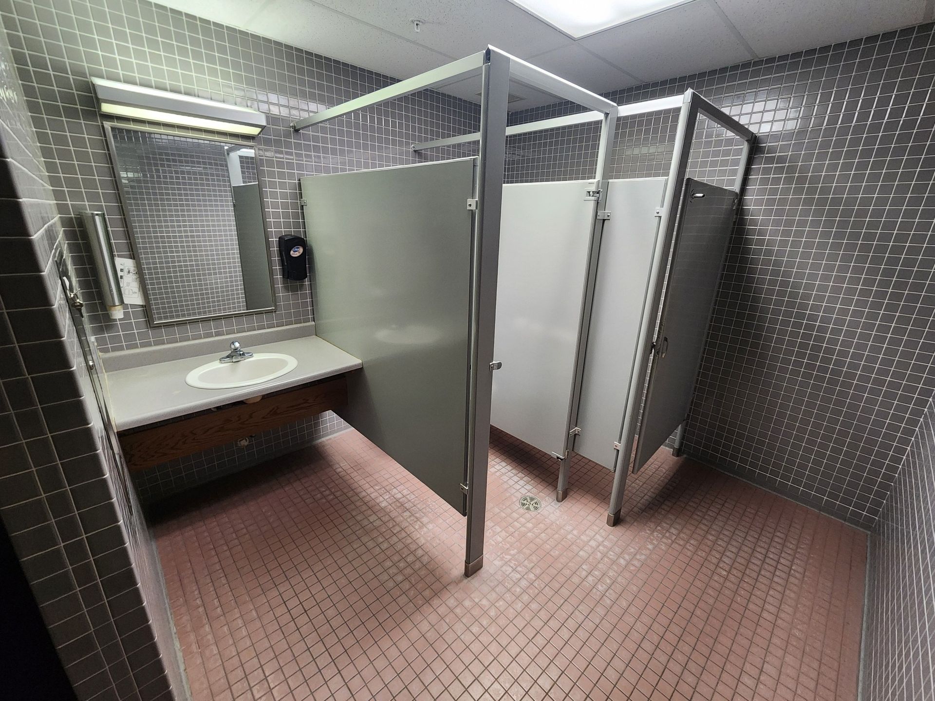 Public restroom with sink, mirror, and gray stall partitions in a tiled room