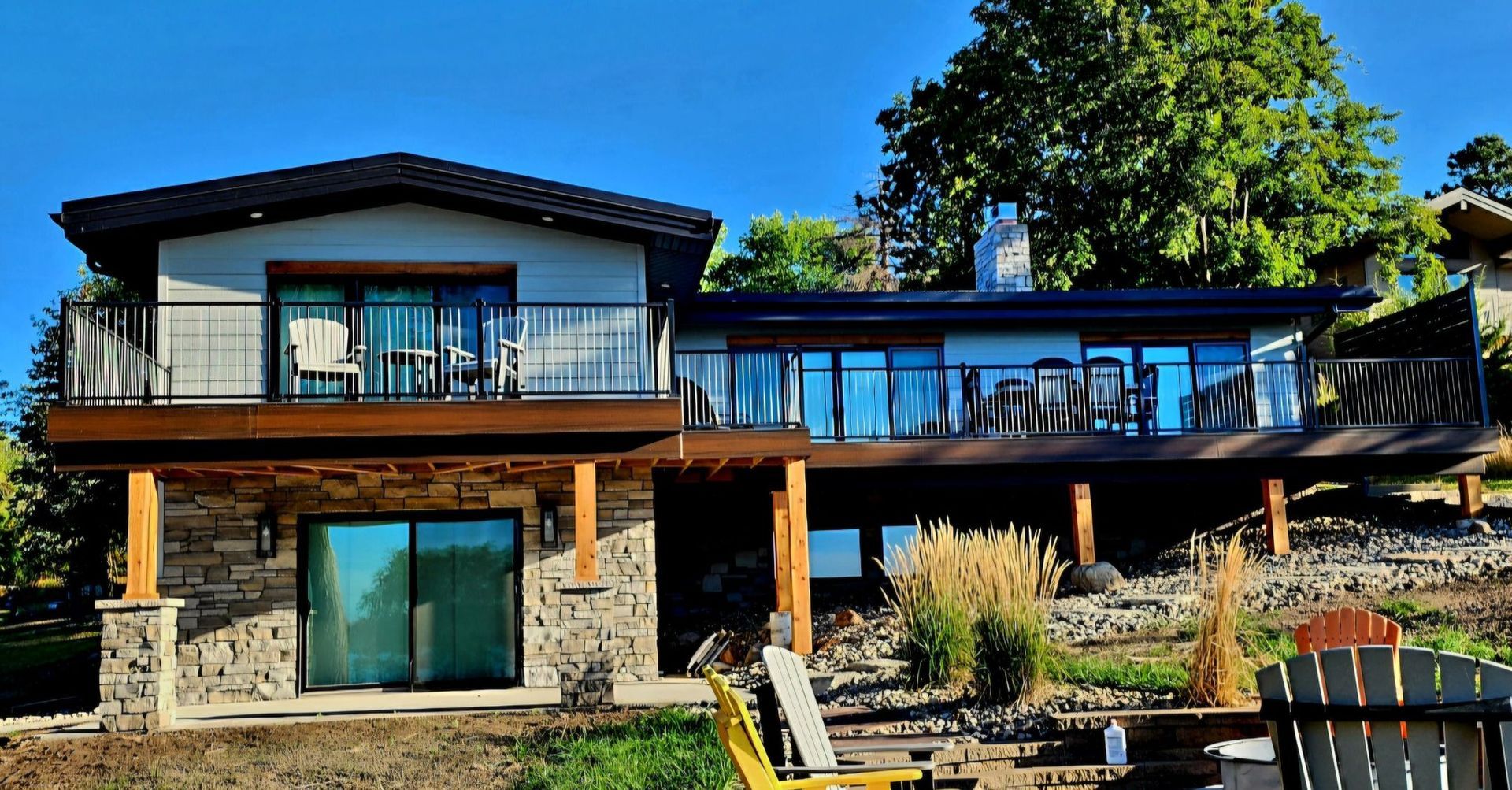 Modern two-story house with stone and wood exterior, large windows, and a sunny deck overlooking a yard