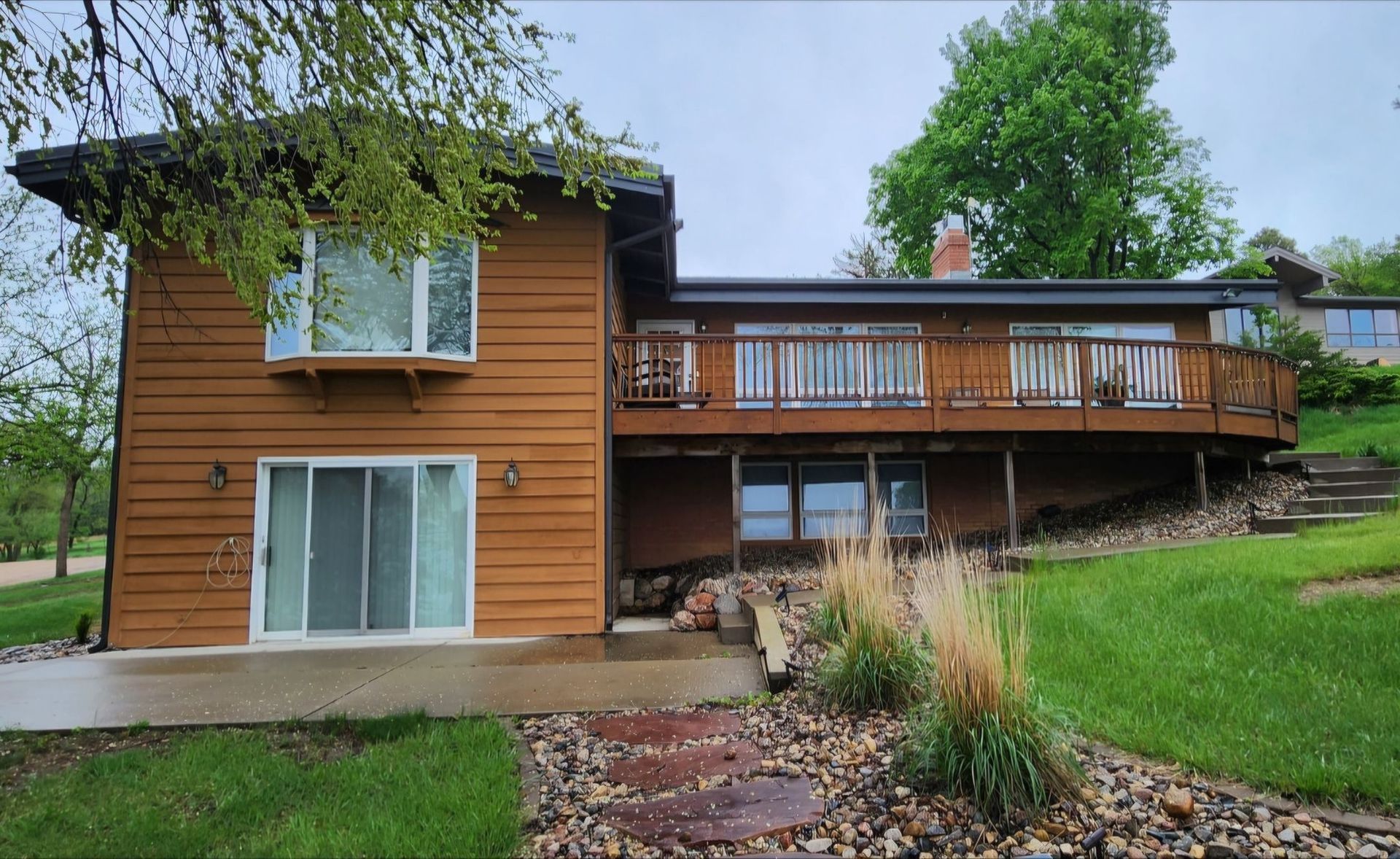 Wood-sided house with a balcony, large windows, and a green yard under cloudy skies.